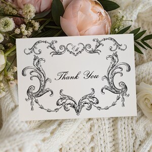 Puede incluir: Una tarjeta crema con las palabras "Thank You" en escritura elegante, enmarcada por adornos ornamentados en blanco y negro. La tarjeta descansa sobre una manta blanca de punto, rodeada de flores rosas y blancas.