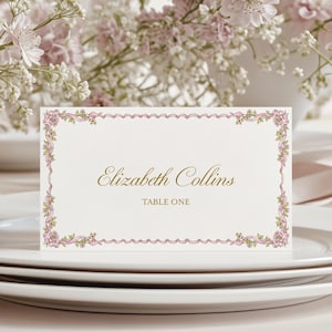 May include: A rectangular white place card featuring a decorative border of pink ribbons and small green leaves. The text Elizabeth Collins is printed in gold script, with the words TABLE ONE printed in gold block letters below the name.