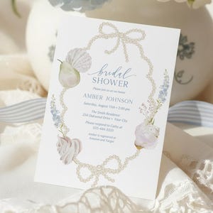 May include: A bridal shower invitation with a pearl and floral design. The invitation features the text "Bridal Shower" and event details. Soft blue and white hydrangeas are in a white vase in the background, with blue and white striped ribbon.