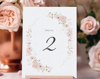 May include: A white table number card with the number 2 in black, surrounded by a floral wreath. The card is displayed on a small wooden easel. Soft pink roses and a copper vase are in the background.