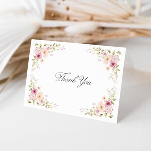 Puede incluir: Una tarjeta blanca con las palabras "Thank You" en escritura elegante, enmarcada por arreglos florales de acuarela en tonos rosa, melocotón y verde. La tarjeta se encuentra sobre un fondo suave y neutro con acentos florales secos.