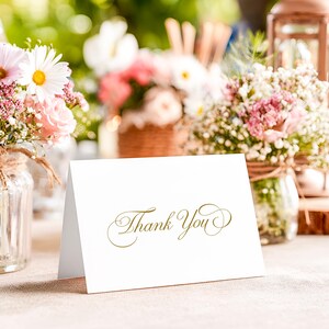 Puede incluir: Una tarjeta blanca plegada con las palabras Thank You impresas en una fuente cursiva dorada. La tarjeta está colocada en posición vertical sobre una superficie plana, con letras elegantes y adornos decorativos al principio y al final.