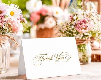 May include: A white folded card with the words Thank You printed in a gold cursive script font. The card is positioned upright on a flat surface, featuring elegant lettering with decorative flourishes at the beginning and end of the text.