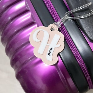 May include: A pink luggage tag with a white letter H and the name Hazel written in white on the tag. The tag is attached to a purple suitcase.