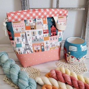 May include: A fabric project bag with a Parisian theme, featuring illustrations of the Eiffel Tower and cafes. The bag is paired with blue and orange yarn skeins and a blue camper van mug. The bag has a gingham top and a dotted bottom.