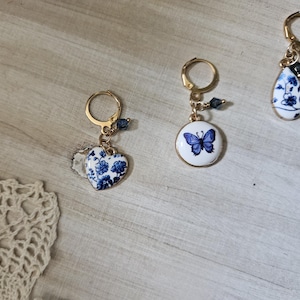 May include: Three gold-toned earrings with blue and white porcelain charms. One earring features a heart-shaped charm with a floral pattern, another has a round charm with a butterfly, and the third has a teardrop-shaped charm.