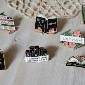 May include: A selection of enamel pins with book-related designs. The pins feature phrases such as "Cool People Read" and "I Cannot Live Without Books." The pins are in various colours and depict open books and stacks of books.
