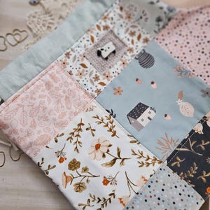 May include: A patchwork item with floral and geometric patterns in pastel shades. The design features squares with flowers, leaves, and a small house. A small panda bear button is sewn onto one of the squares.
