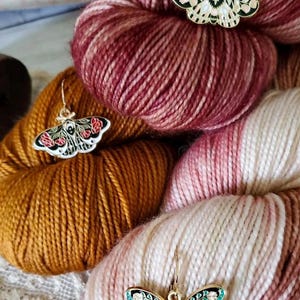 May include: Three skeins of yarn in different colours: a deep red, a brown, and a white with pink and red streaks. Each skein has a gold-coloured metal butterfly charm attached to it. The butterflies have black and white wings with floral details and a green accent.