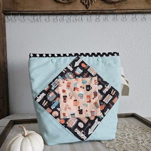 May include: A light blue and white gingham fabric bag featuring a central diamond-shaped panel with a black background printed with trucks, pumpkins, and coffee cups, and a smaller peach-colored square pocket with a coffee cup and leaf pattern.
