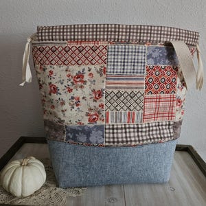 Puede incluir: Un bolso de tela de patchwork con una variedad de patrones que incluyen estampados florales, diseños geométricos rojos y blancos, y cuadros vichy marrones. El bolso tiene una base de mezclilla azul, un borde superior vichy y cordones.