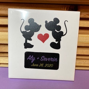 May include: White ceramic tile with a black silhouette of Mickey and Minnie Mouse kissing. A red heart is between them. A black rectangle with the text "Aly + Severin June 26, 2020" is at the bottom.