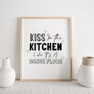 May include: A framed print with the text "Kiss in the kitchen like it's a dance floor" in black text on a white background. The frame is light wood.