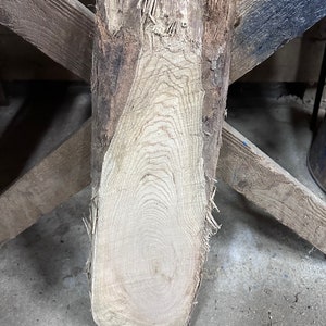 May include: A close-up of a rough-hewn, light brown wood slab with a visible grain pattern. The wood has a natural, rustic look and is likely a piece of lumber.