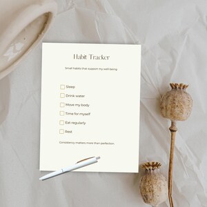 Op de afbeelding: Een crèmekleurige habit tracker met de titel "Habit Tracker" en een lijst met gewoonten: Slaap, Drink water, Beweeg mijn lichaam, Tijd voor mezelf, Eet regelmatig en Rust. Een pen ligt op de pagina. Gedroogde zaaddozen staan op de achtergrond.