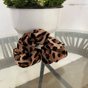 May include: A leopard print scrunchie in brown and black tones. The hair accessory is made of soft fabric and is placed on a glass surface. A white pot with greenery is in the background.