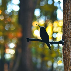 May include: A black metal hummingbird silhouette perched on a branch, mounted on a tree trunk. The background is a blurred out-of-focus image of a forest with bokeh.