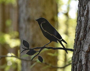 May include: A black metal silhouette of a bird perched on a branch with leaves, mounted on a tree trunk.