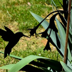 May include: A black metal hummingbird silhouette in flight, approaching a stylized flower silhouette. The artwork is set against a backdrop of green grass and foliage, creating a contrast of light and shadow. The design is suitable for garden decor.