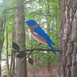 May include: A bluebird metal sculpture perched on a metal branch with leaves. The sculpture is attached to a tree trunk in a forest setting.