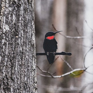 May include: A black metal hummingbird with a red throat perched on a branch in a forest setting.