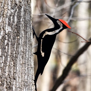 May include: Black metal silhouette of a woodpecker perched on a tree trunk. The woodpecker has a red crest and white markings on its black body.