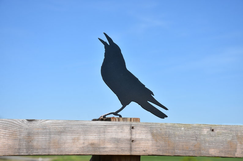May include: Black metal silhouette of a crow perched on a wooden fence post against a blue sky.