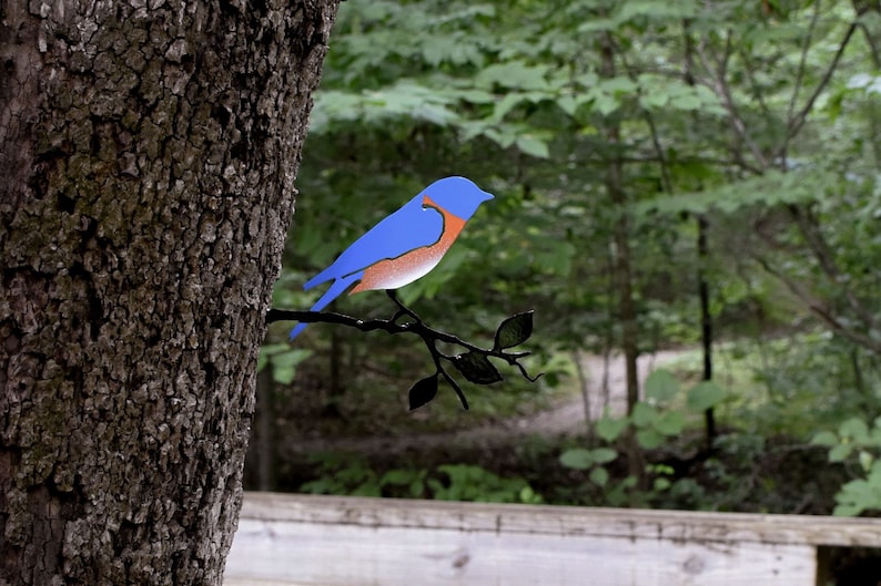 May include: A bluebird metal silhouette perched on a black metal branch with leaves. The bird is painted blue with an orange breast. The silhouette is attached to a tree trunk.