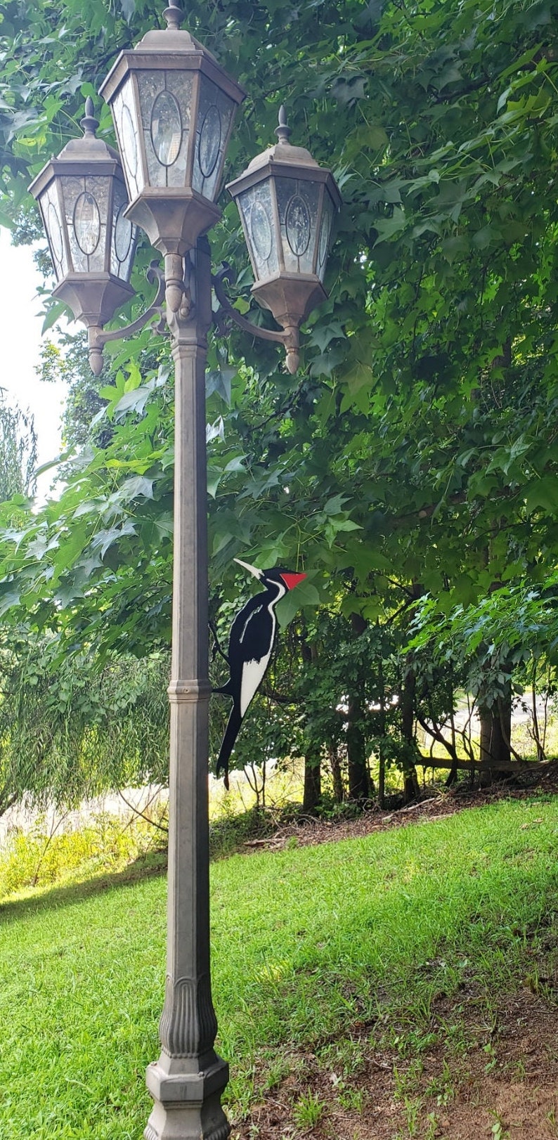 May include: A black and white woodpecker with a red crest is perched on a lamppost with three black lanterns. The lamppost is in a grassy area with green trees in the background.