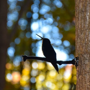 May include: A black metal hummingbird silhouette mounted on a tree trunk. The hummingbird is perched on a branch with its wings spread out.