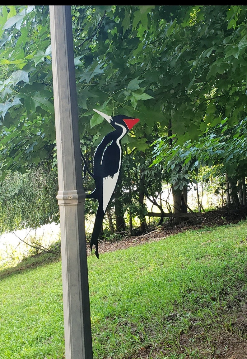 May include: Black and white metal woodpecker silhouette with a red beak mounted on a gray pole. The woodpecker is facing right and is perched on a branch. The background is a green lawn and trees.