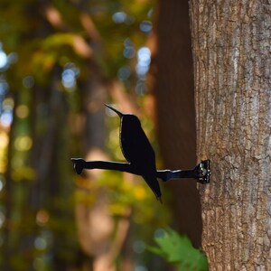 May include: A black metal hummingbird silhouette mounted on a tree trunk. The hummingbird is perched on a branch with its wings spread out.