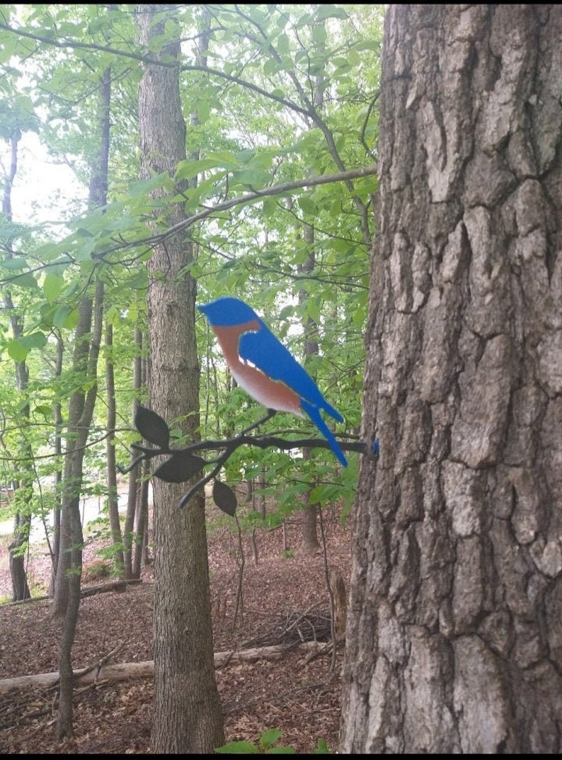 May include: A bluebird metal sculpture perched on a metal branch with leaves. The sculpture is attached to a tree trunk in a forest setting.