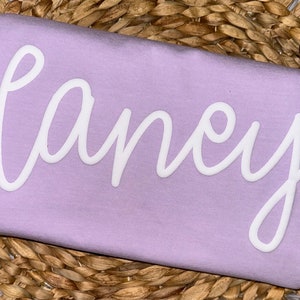 May include: A light purple t-shirt with the name "Laney" in white cursive lettering. The shirt is displayed on a woven, light brown surface.