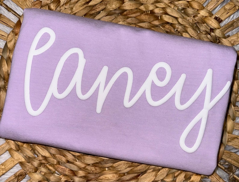 May include: A light purple t-shirt with the name "Laney" in white cursive lettering. The shirt is displayed on a woven, light brown surface.