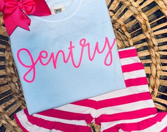 May include: A light blue t-shirt with the name "Gentry" written in pink script. The shirt is paired with white and pink striped shorts with a ruffle trim.