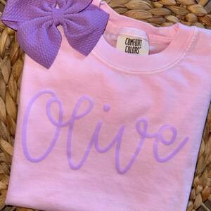 May include: A light pink t-shirt with the name "Olive" in a light purple script. A large purple bow is placed at the top left. The shirt has a Comfort Colors tag.