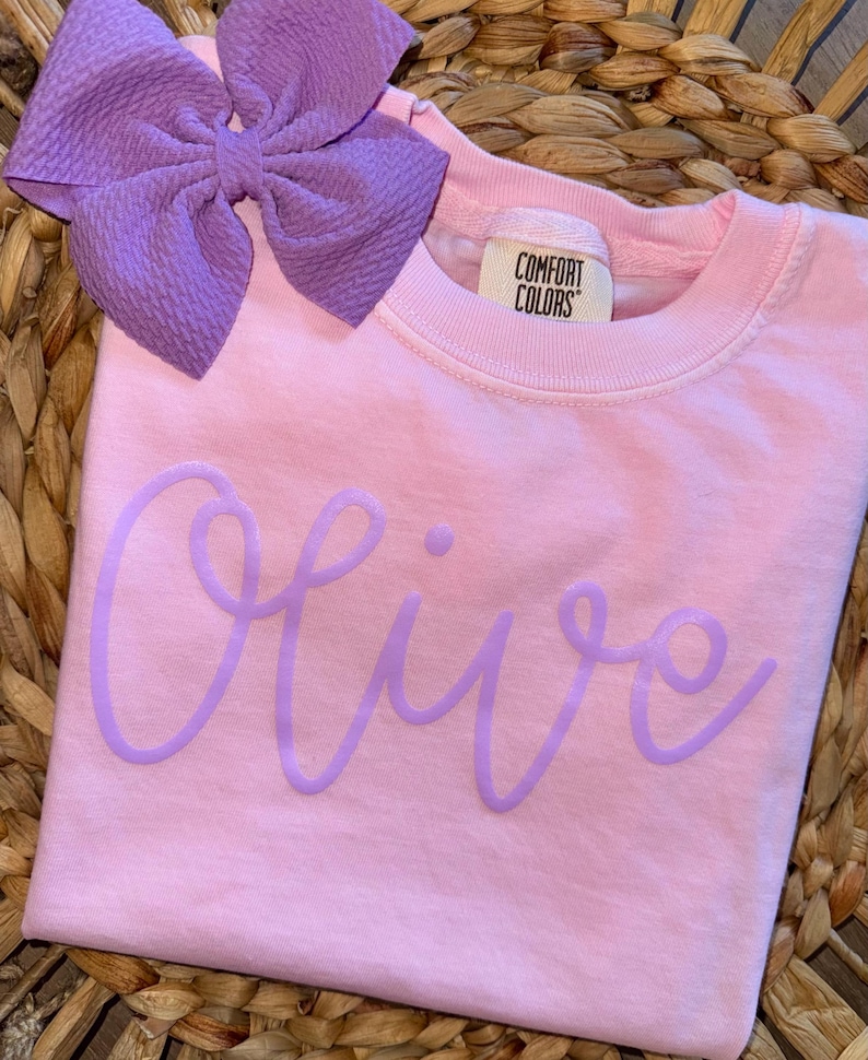 May include: A light pink t-shirt with the name "Olive" in a light purple script. A large purple bow is placed at the top left. The shirt has a Comfort Colors tag.