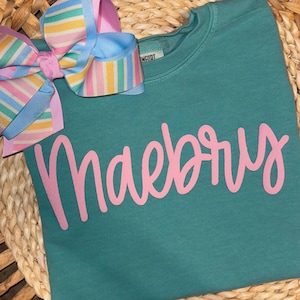 May include: Teal sweatshirt with the name "Maebry" in pink script. A large, colorful bow with pastel stripes and pink accents is attached to the top left. The bow has blue, yellow, and pink stripes.