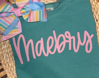 May include: Teal sweatshirt with the name "Maebry" in pink script. A large, colorful bow with pastel stripes and pink accents is attached to the top left. The bow has blue, yellow, and pink stripes.