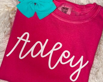 May include: A pink sweatshirt with the name "Adley" written in white cursive lettering. The sweatshirt has a small tag that says "Comfort Colors".