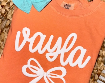 May include: A bright orange t-shirt with the name "vayla" written in white cursive lettering. A white bow is below the name. The shirt has a tag that says "Comfort Colors".