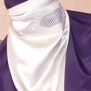 Puede incluir: Un niqab de satén en blanco y morado oscuro. La parte blanca presenta un diseño bordado y una cobertura para los ojos de malla. La tela se drapea suavemente, creando una apariencia modesta y elegante.