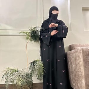 May include: A black abaya with pink bow embroidery. The abaya features wide sleeves and is worn with a matching head covering and face veil. The person is wearing white sandals with floral embellishments. A decorative plant in a textured pot is in the foreground.