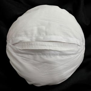 Könnte beinhalten: Ein weißes, rundes Kissen mit einer eingenähten, rechteckigen Öffnung. Das Kissen ist aus einem weichen Stoff, möglicherweise Baumwolle. Das Kissen befindet sich auf einem schwarzen Hintergrund.