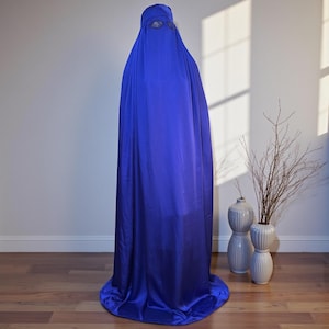 May include: A full-length, royal blue burqa. The garment drapes from head to floor, with a small, eye-level opening. The fabric has a slight sheen. The background is a plain white wall with a vase of branches.
