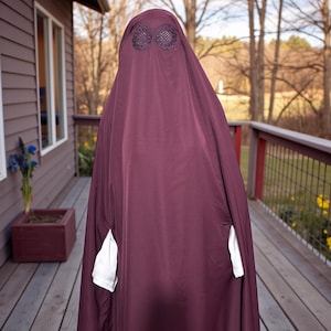 May include: A burgundy-colored burqa with a decorative eye covering. The garment has white pockets and is worn outdoors. The background includes a wooden deck, a house, and trees.