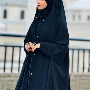 Op de afbeelding: Een zwarte abaya met een lange, vloeiende rok en lange mouwen. De abaya heeft een knoopsluiting aan de voorkant en een hoge kraag. De vrouw draagt een zwarte hijab en een zonnebril.