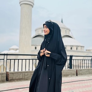 Op de afbeelding: Een vrouw draagt een zwarte abaya met een lange, vloeiende rok en een zwarte hoofddoek. Ze staat voor een wit gebouw met een hoge minaret.