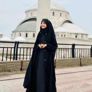 Op de afbeelding: Een vrouw draagt een zwarte abaya met een lange, vloeiende rok en een zwarte hijab. Ze staat voor een moskee met een witte koepel.